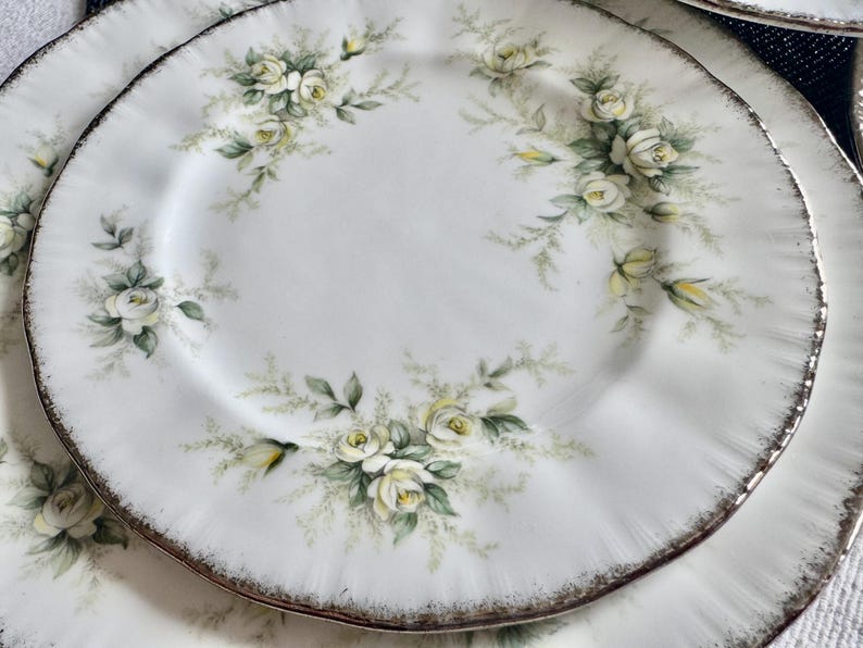 May include: A set of white porcelain plates with a delicate floral design featuring white roses and green leaves. The plates have a gold rim and a scalloped edge.