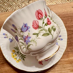 May include: A white teacup and saucer with a floral pattern. The pattern features pink, blue, and green flowers on a white background. The teacup has a gold rim.