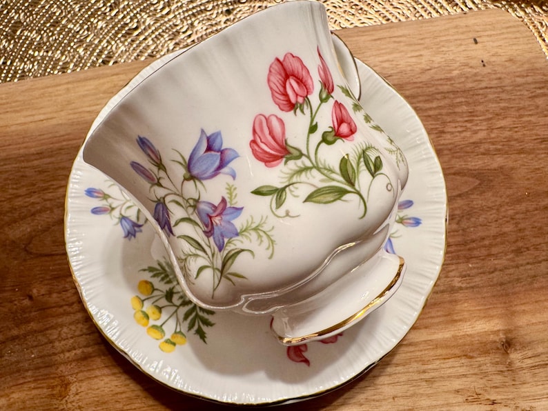 May include: A white teacup and saucer with a floral pattern. The pattern features pink, blue, and green flowers on a white background. The teacup has a gold rim.