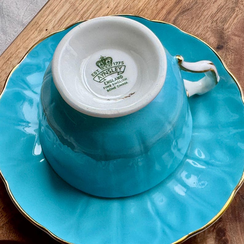 May include: A turquoise teacup and saucer set with gold trim. The teacup has a white interior with the words "EST. 1775 AYNSLEY ENGLAND FINE ENGLISH BONE CHINA" printed on the bottom.