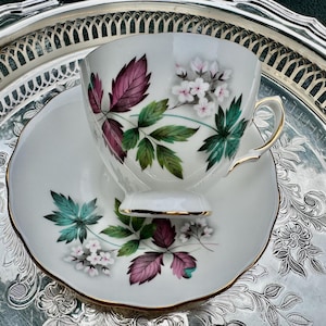 May include: White teacup and saucer set with a floral design featuring burgundy, green, and teal leaves and small white flowers. The cup and saucer have gold trim. The set is displayed on a silver serving tray.