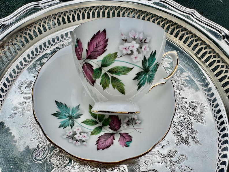 May include: White teacup and saucer set with a floral design featuring burgundy, green, and teal leaves and small white flowers. The cup and saucer have gold trim. The set is displayed on a silver serving tray.