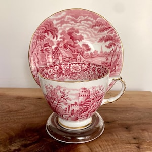 Pu&ograve; includere: Un set vintage di tazza e piattino da t&egrave; con una scena pastorale rossa e bianca. La tazza da t&egrave; presenta un disegno di castello, mentre il piattino mostra un paesaggio. Entrambi i pezzi hanno finiture dorate. La tazza da t&egrave; poggia su un piattino di vetro trasparente.