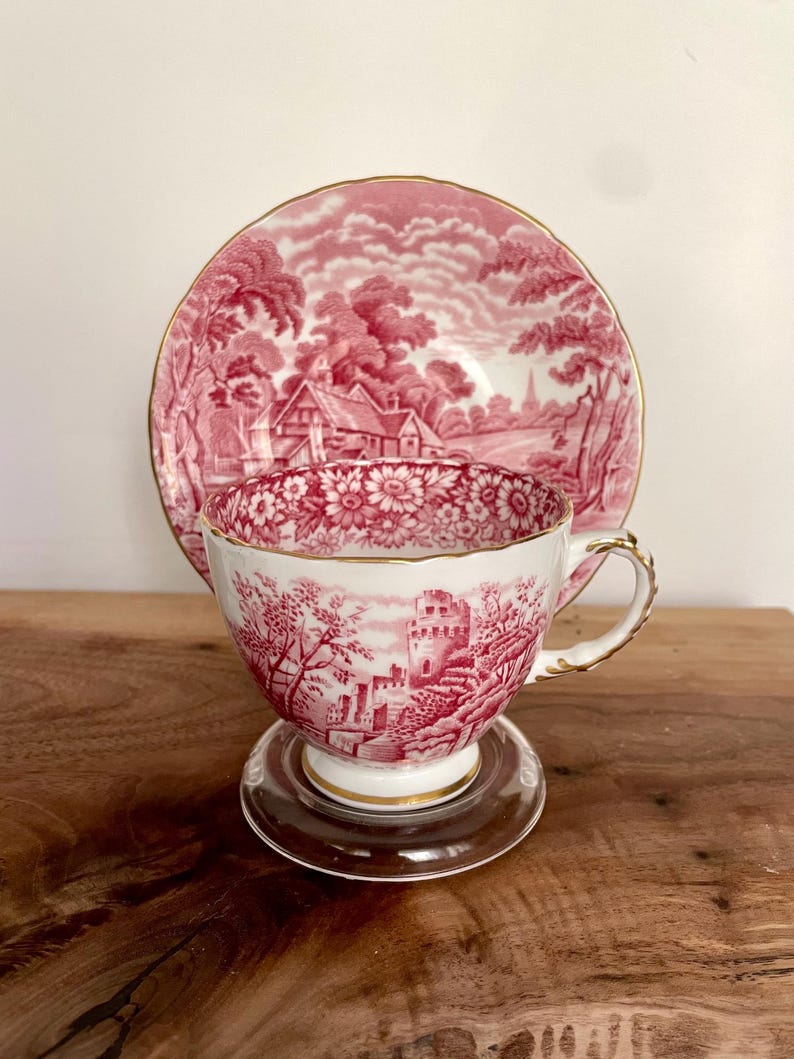 Pu&ograve; includere: Un set vintage di tazza e piattino da t&egrave; con una scena pastorale rossa e bianca. La tazza da t&egrave; presenta un disegno di castello, mentre il piattino mostra un paesaggio. Entrambi i pezzi hanno finiture dorate. La tazza da t&egrave; poggia su un piattino di vetro trasparente.