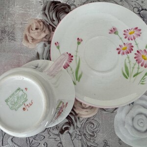 May include: A vintage white teacup, saucer, and plate set. The set features a floral design with pink and yellow daisies and green stems. The teacup has a pink handle and the set is marked "Shelley England".