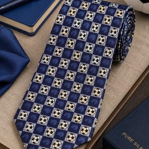 May include: A necktie featuring a repeating checkerboard pattern of dark blue and light gray squares. Each light gray square contains a small black and white soccer ball graphic. The tie is made of pure silk and has a pointed end.