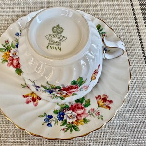 Puede incluir: Una taza de t&eacute; y un platillo blancos con un patr&oacute;n floral. La taza de t&eacute; tiene un borde dorado y el platillo tiene un borde festoneado. La taza de t&eacute; tiene las palabras "Aynsley England Bone China" impresas en la parte inferior.