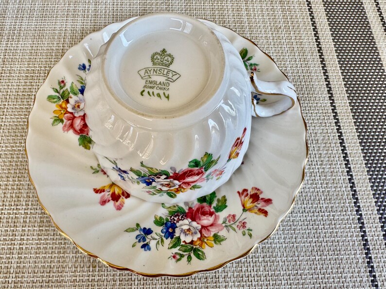 Puede incluir: Una taza de t&eacute; y un platillo blancos con un patr&oacute;n floral. La taza de t&eacute; tiene un borde dorado y el platillo tiene un borde festoneado. La taza de t&eacute; tiene las palabras "Aynsley England Bone China" impresas en la parte inferior.