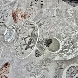 May include: A clear glass candy dish with a matching lid, featuring a pressed diamond and starburst pattern. The bowl has a scalloped rim, and the lid includes a faceted knob handle. The glass is transparent with a reflective, textured surface.