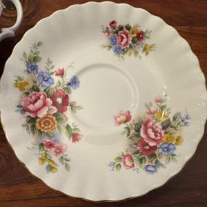 May include: A white porcelain saucer with a floral design. The saucer features a delicate scalloped edge and a central well for a teacup. The floral design includes pink, red, yellow, blue, and green flowers.