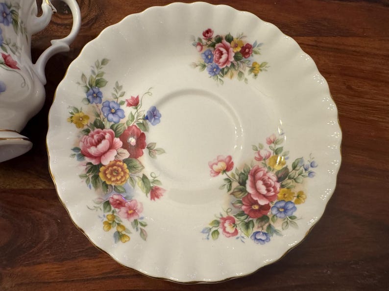 May include: A white porcelain saucer with a floral design. The saucer features a delicate scalloped edge and a central well for a teacup. The floral design includes pink, red, yellow, blue, and green flowers.