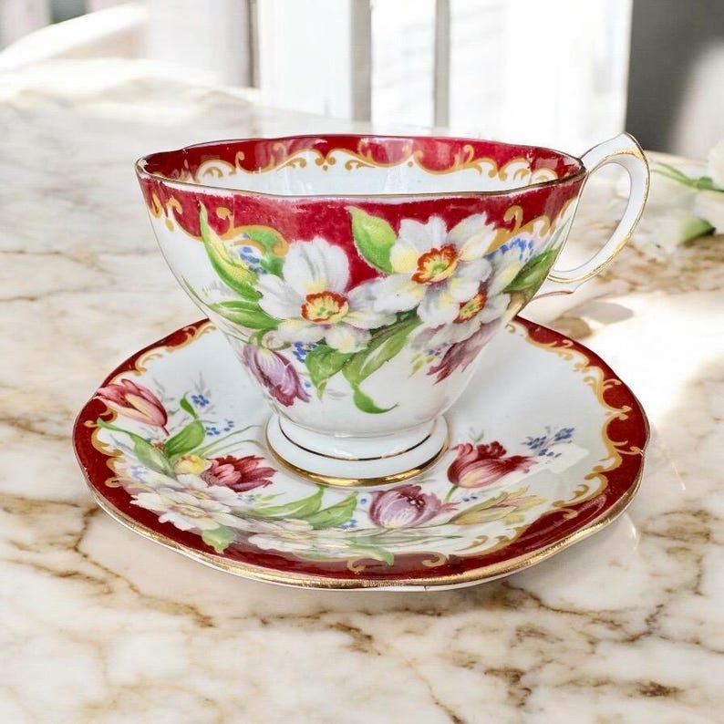 May include: A vintage teacup and saucer set. The teacup and saucer are white with a red border and gold trim. The teacup and saucer are decorated with a floral pattern of white, yellow, and purple flowers.
