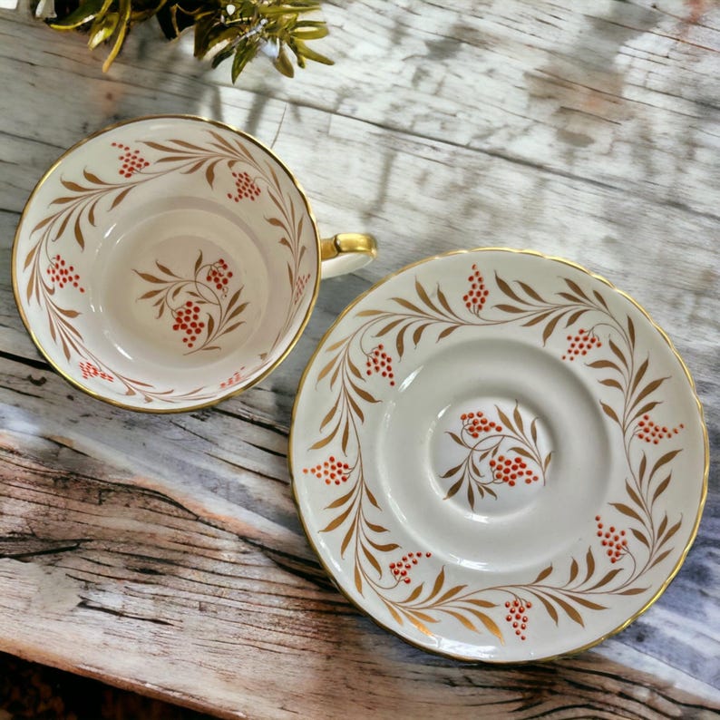 Pu&ograve; includere: Un set vintage di tazza e piattino da t&egrave;. La tazza e il piattino in porcellana bianca presentano un bordo dorato e un motivo di foglie dorate e bacche rosse. La tazza ha un manico dorato. Il set &egrave; su una superficie di legno.