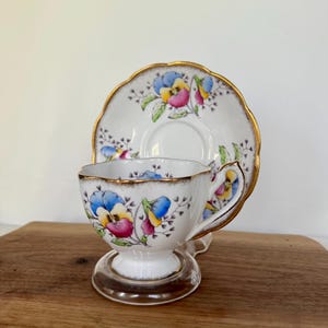 May include: A white teacup and saucer set with a floral design featuring blue, pink, and yellow flowers. The teacup and saucer have gold trim. The teacup is on a clear, round stand. The set is on a wooden surface.