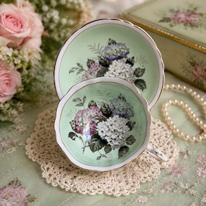 May include: A pale mint green porcelain teacup and matching saucer set featuring a scalloped edge with gold trim and a detailed floral print of white, blue, and purple hydrangea blossoms with dark green leaves centered on the interior and saucer.