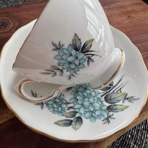May include: A white teacup and saucer with a gold trim. The teacup and saucer are decorated with a floral pattern of blue and green flowers.