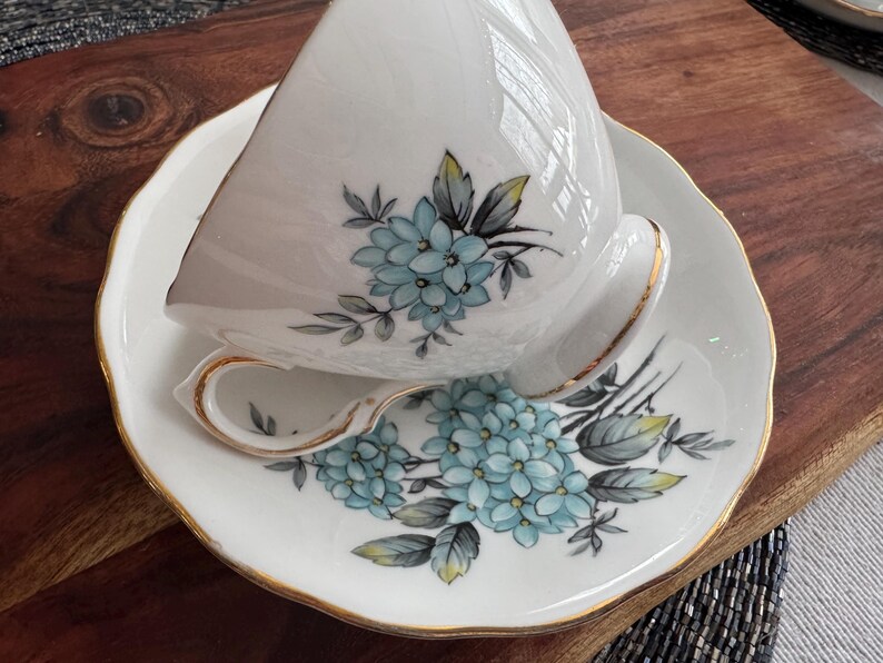 May include: A white teacup and saucer with a gold trim. The teacup and saucer are decorated with a floral pattern of blue and green flowers.