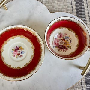 May include: A red and gold teacup and saucer set with a floral pattern. The cup and saucer have a white background with a red rim and gold trim. The floral pattern is in shades of pink, yellow, and purple.