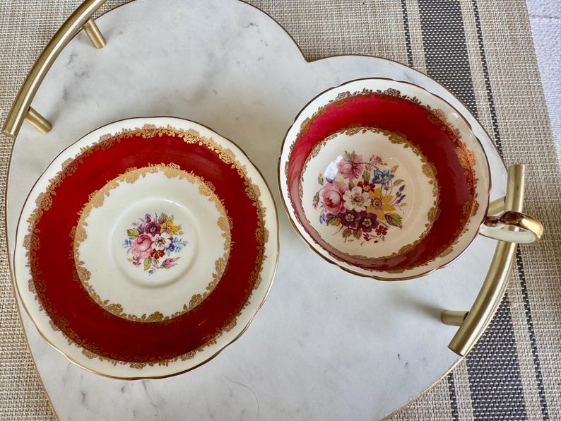May include: A red and gold teacup and saucer set with a floral pattern. The cup and saucer have a white background with a red rim and gold trim. The floral pattern is in shades of pink, yellow, and purple.