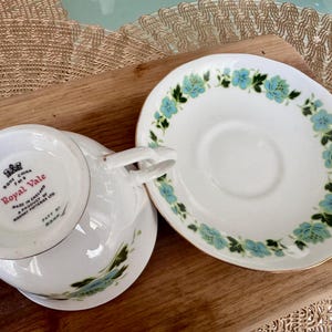 May include: A white teacup and saucer with a blue and green floral pattern. The teacup has the words "Royal Vale" printed on the bottom. The saucer has a gold rim.