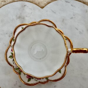Puede incluir: Juego de taza y platillo blancos con ribete dorado y detalles florales. La taza y el platillo tienen un borde festoneado. El juego est&aacute; colocado sobre una bandeja de m&aacute;rmol en forma de coraz&oacute;n con asas doradas.