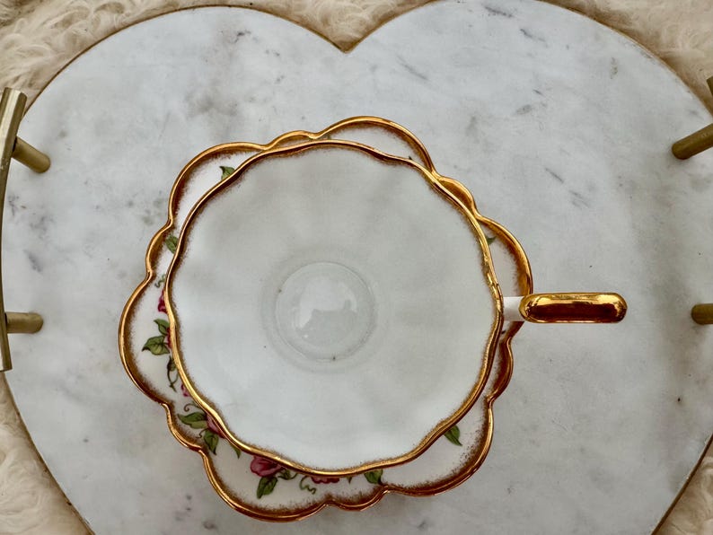 Puede incluir: Juego de taza y platillo blancos con ribete dorado y detalles florales. La taza y el platillo tienen un borde festoneado. El juego est&aacute; colocado sobre una bandeja de m&aacute;rmol en forma de coraz&oacute;n con asas doradas.