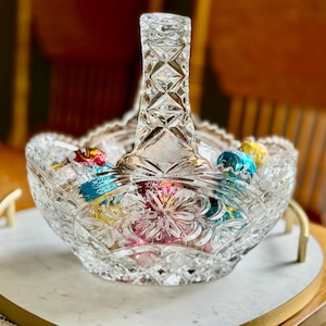 May include: A clear, cut-glass basket with a handle, filled with colorful wrapped candies. The basket has a diamond and floral pattern. The handle is also cut glass. The candies are in shades of blue, yellow, pink, and red.