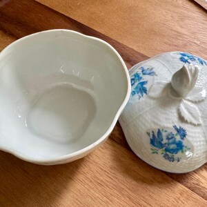 May include: A white ceramic bowl with a matching lid. The lid is shaped like a fruit with a stem and leaf detail. Blue floral patterns decorate the lid. The bowl is open, revealing its interior.
