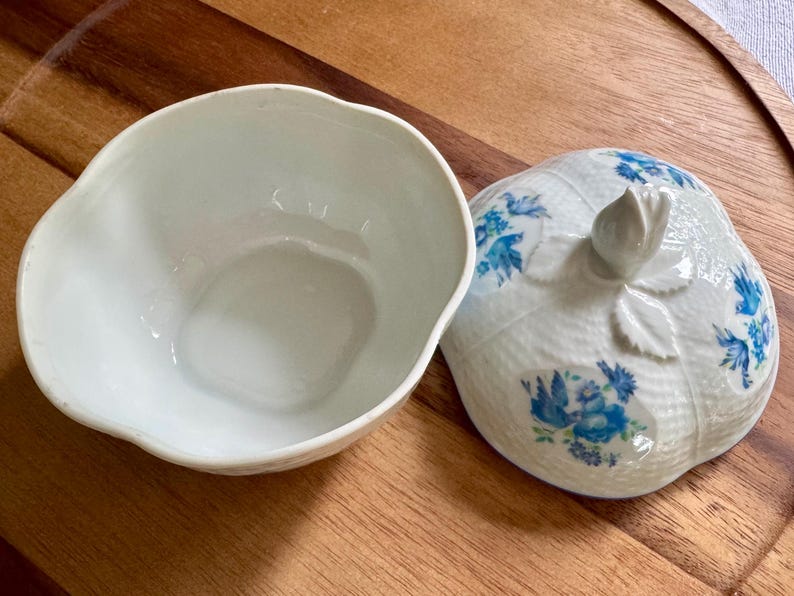 May include: A white ceramic bowl with a matching lid. The lid is shaped like a fruit with a stem and leaf detail. Blue floral patterns decorate the lid. The bowl is open, revealing its interior.