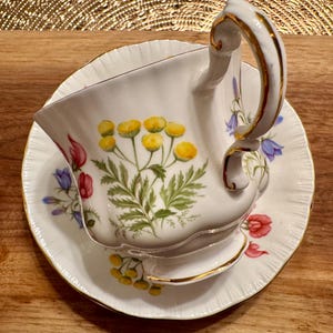May include: A white teacup and saucer with a floral pattern. The cup has a gold rim and a delicate handle. The saucer has a scalloped edge and a floral pattern.