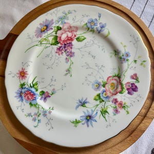 May include: White ceramic plate with a floral design featuring pink, blue, and purple flowers. The plate has a scalloped edge and is set on a wooden serving tray. The floral pattern includes roses, daisies, and other wildflowers.