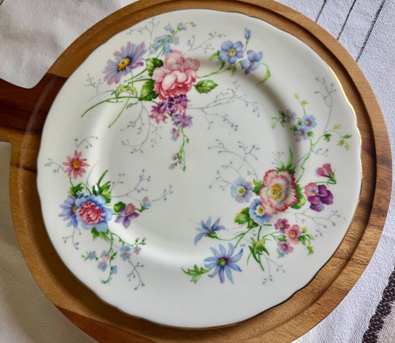 May include: White ceramic plate with a floral design featuring pink, blue, and purple flowers. The plate has a scalloped edge and is set on a wooden serving tray. The floral pattern includes roses, daisies, and other wildflowers.