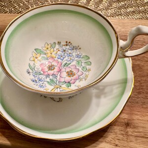 May include: A vintage teacup and saucer set with a floral design. The cup has a green rim and a gold trim. The saucer has a green rim and a gold trim. The floral design features pink, white, and yellow flowers.