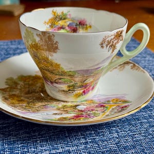May include: A white porcelain teacup and saucer set featuring a colorful landscape scene with a stone bridge, trees, and flowers. The teacup has a pale green handle and a scalloped rim, while both pieces are finished with a thin gold trim.