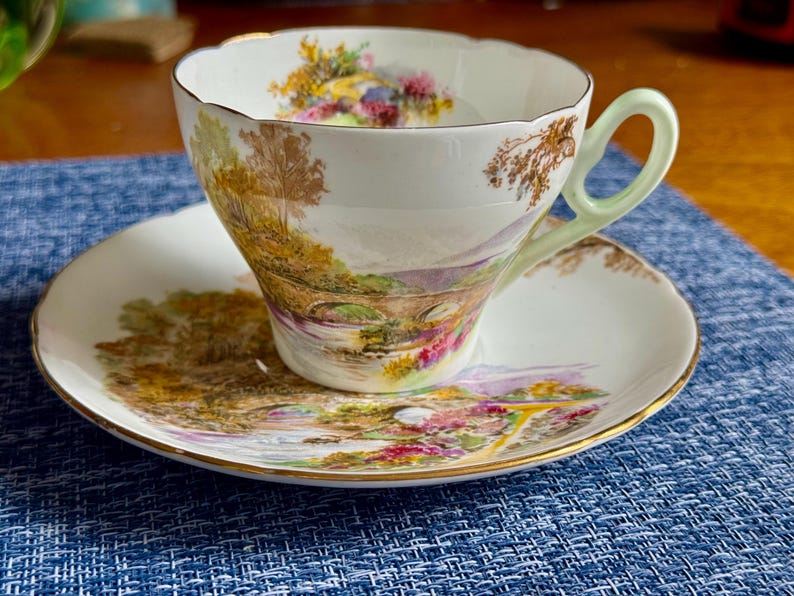 May include: A white porcelain teacup and saucer set featuring a colorful landscape scene with a stone bridge, trees, and flowers. The teacup has a pale green handle and a scalloped rim, while both pieces are finished with a thin gold trim.