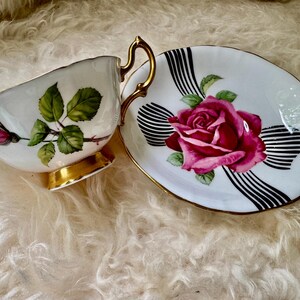 May include: A white teacup and saucer set with a floral rose design. The teacup has a gold handle and base, and the saucer features a large pink rose with black and white stripes. The set is a vintage style.