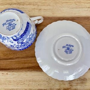 May include: A vintage teacup and saucer set with a blue and white floral design. The teacup has a gold handle and the saucer has a scalloped edge. The set is marked "Blue Rhapsody Gladstone" and "Made in England".