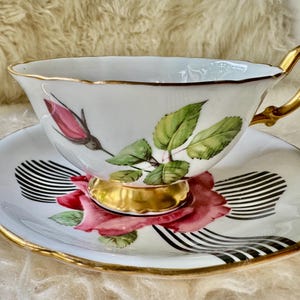 May include: White teacup and saucer set with gold trim. The cup and saucer feature a pink rose and green leaf design. The saucer has a black and white striped pattern. The handle and base of the cup are gold.
