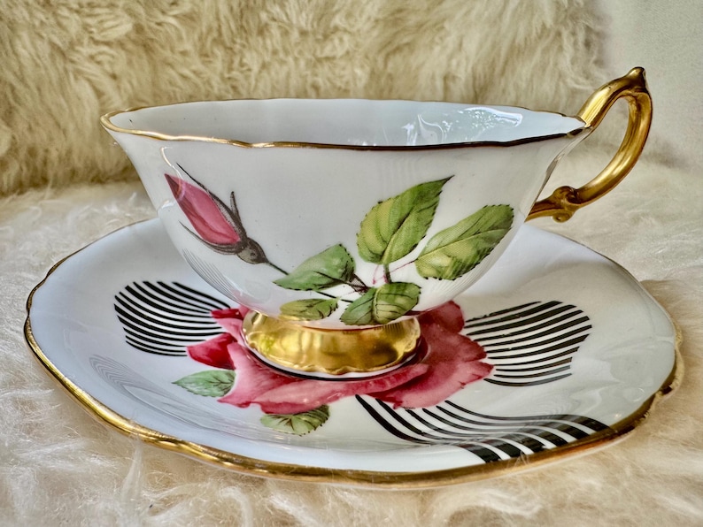 May include: White teacup and saucer set with gold trim. The cup and saucer feature a pink rose and green leaf design. The saucer has a black and white striped pattern. The handle and base of the cup are gold.