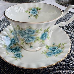 May include: A white teacup and saucer set with a delicate floral pattern featuring blue roses. The set has a gold trim around the edges.