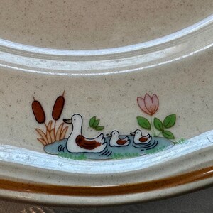 May include: A beige ceramic plate with a brown rim and a duck family design. The design features a mother duck and two ducklings swimming in a pond, with cattails and a pink flower.