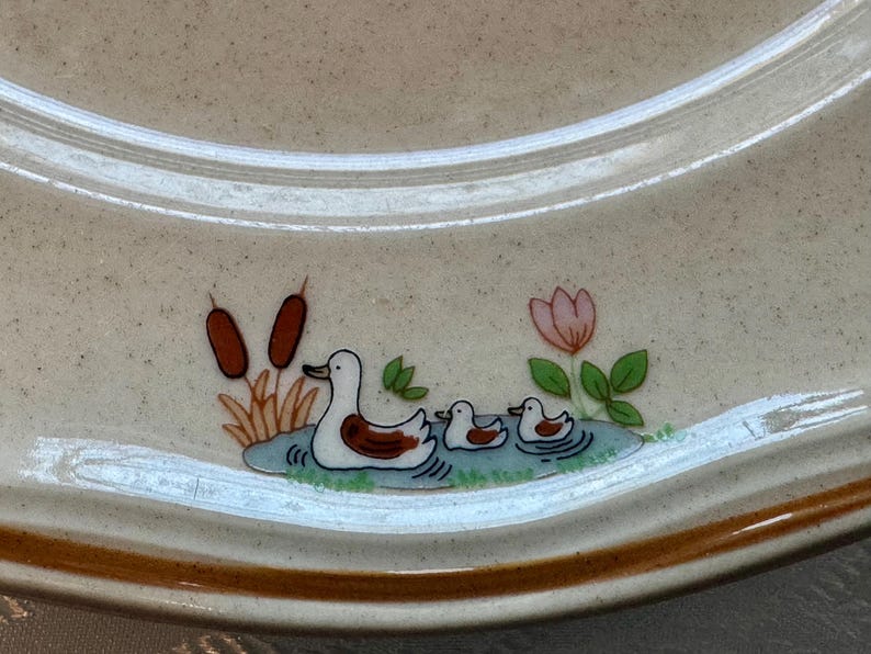 May include: A beige ceramic plate with a brown rim and a duck family design. The design features a mother duck and two ducklings swimming in a pond, with cattails and a pink flower.