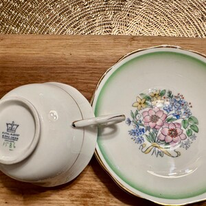 May include: A white teacup and saucer with a green rim and floral design. The saucer has a bouquet of pink, yellow, and blue flowers with green leaves. The teacup has a gold rim and the Royal Albert Bone China England mark on the bottom.