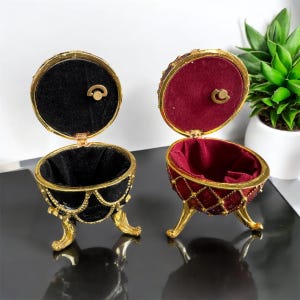 May include: Two ornate gold egg-shaped trinket boxes with red and black velvet interiors. The boxes have a hinged lid and stand on three gold feet.