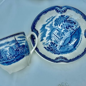 Puede incluir: Un juego de taza y platillo blancos con un dise&ntilde;o azul y blanco. La taza y el platillo presentan una escena de paisaje detallada con una iglesia y &aacute;rboles. El juego es probablemente vintage o antiguo.