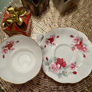 May include: A white teacup and saucer with a floral design featuring pink and red roses. The saucer has a gold rim.
