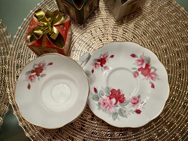 May include: A white teacup and saucer with a floral design featuring pink and red roses. The saucer has a gold rim.