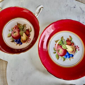 May include: A red teacup and saucer set with a white floral design featuring a pear, apple, and grapes. The set has a gold trim.