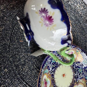 以下が含まれることがあります： 青と金の色の花柄が施された、ヴィンテージの磁器製のティーカップとソーサーのセット。カップは緑色の取っ手、ソーサーは白い中心に花柄が描かれている。