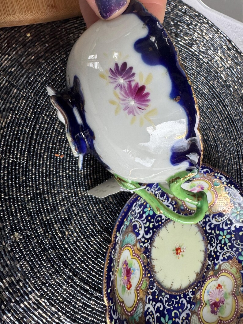 以下が含まれることがあります： 青と金の色の花柄が施された、ヴィンテージの磁器製のティーカップとソーサーのセット。カップは緑色の取っ手、ソーサーは白い中心に花柄が描かれている。